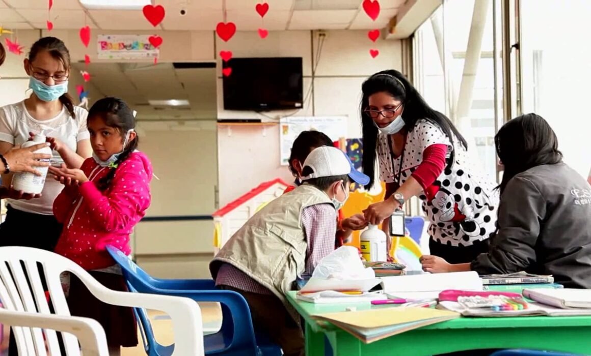 Aulas hospitalarias: niños pequeños en una mesa alrededor de una profesora con tapabocas y elementos de clase
