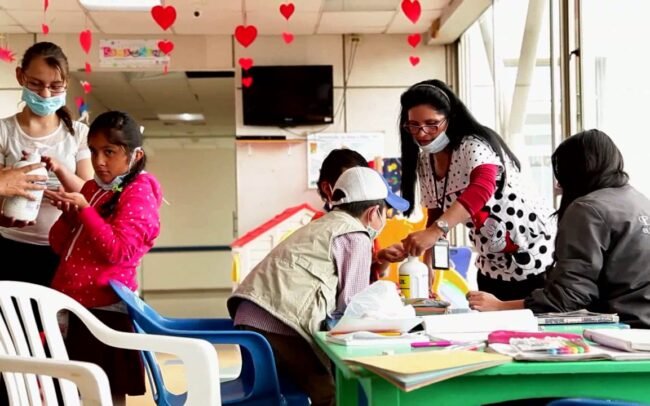 Aulas hospitalarias: niños pequeños en una mesa alrededor de una profesora con tapabocas y elementos de clase